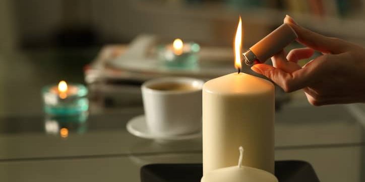 Hand lighting a white candle with a lighter, surrounded by additional candles and a coffee cup, illustrating energy conservation methods during rolling blackouts.
