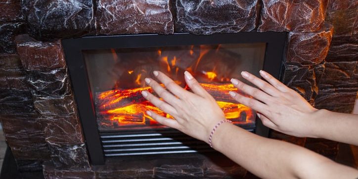 Hands warming near an electric fireplace with glowing logs, emphasizing safety and comfort in home heating solutions.