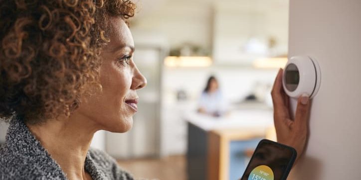 Woman adjusting a smart thermostat while holding a smartphone, illustrating energy-saving technology in a modern home setting.