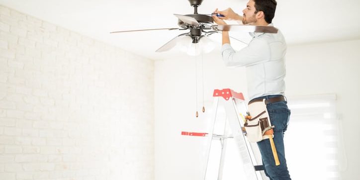 Man installing a ceiling fan on a ladder in a bright indoor setting, demonstrating ceiling fan installation services relevant to Cathey Electric's buying guide.