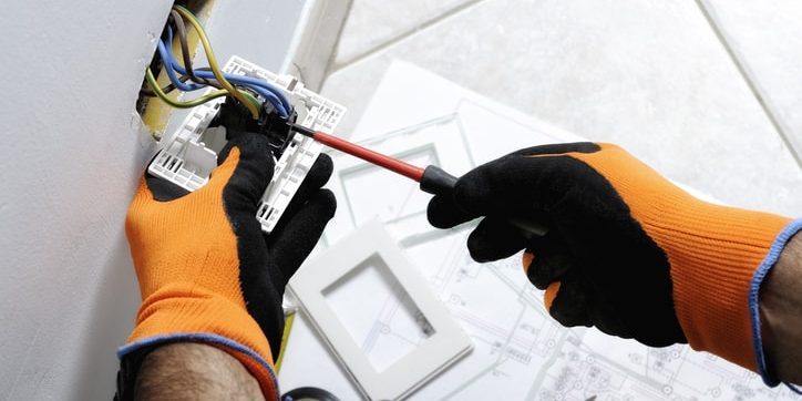 Electrician wearing orange gloves working on electrical wiring installation, screwdriver in hand, with blue and yellow wires visible, surrounded by tools and installation plans.