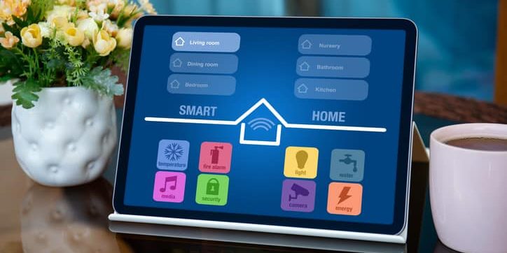 Tablet displaying a smart home automation interface with controls for temperature, fire alarms, lighting, media, security, cameras, and energy management, alongside a cup of coffee and a potted flower arrangement.